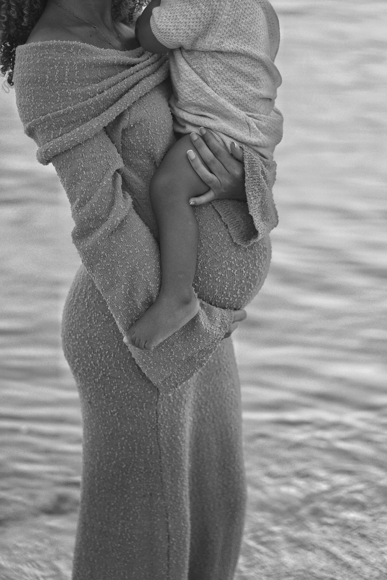 A toddler in cream knitwear holding their mother's hand while walking in the shallow water at sunset