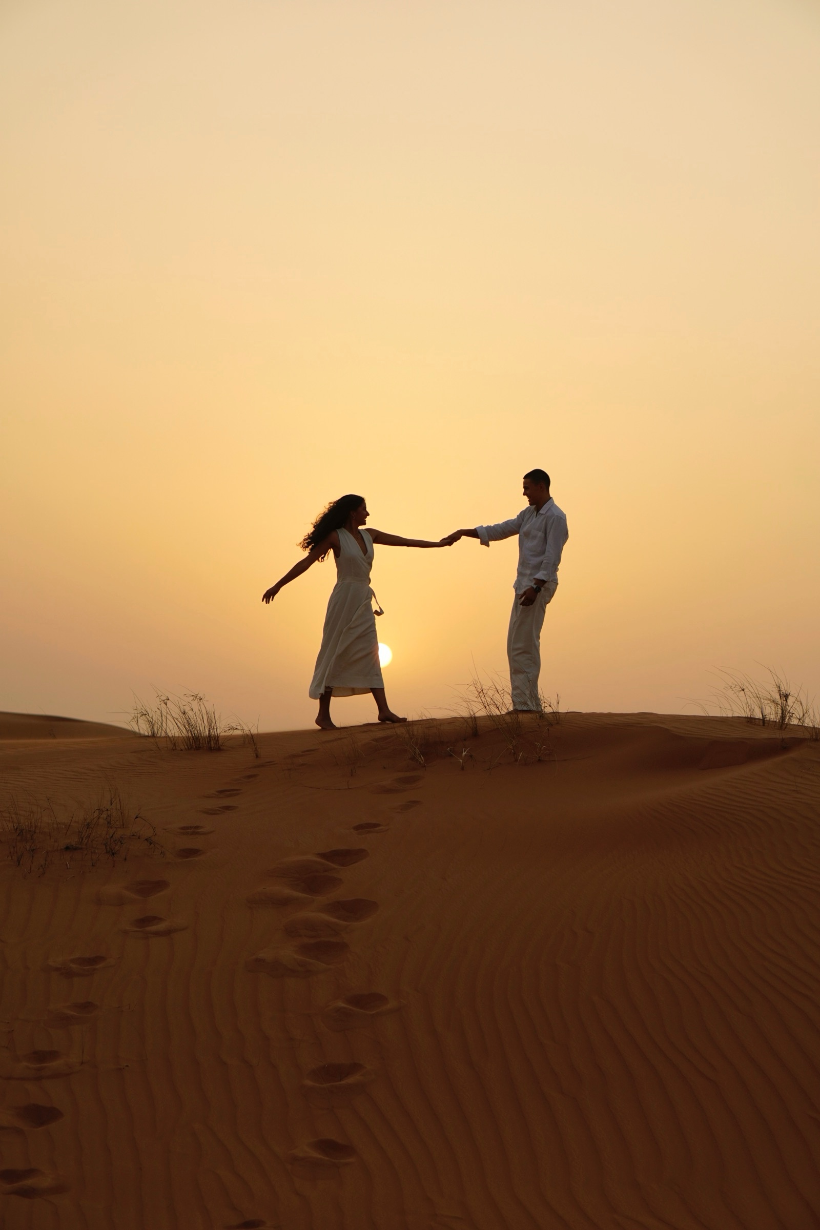 Couple holding hands, silhouetted on a dune at sunset