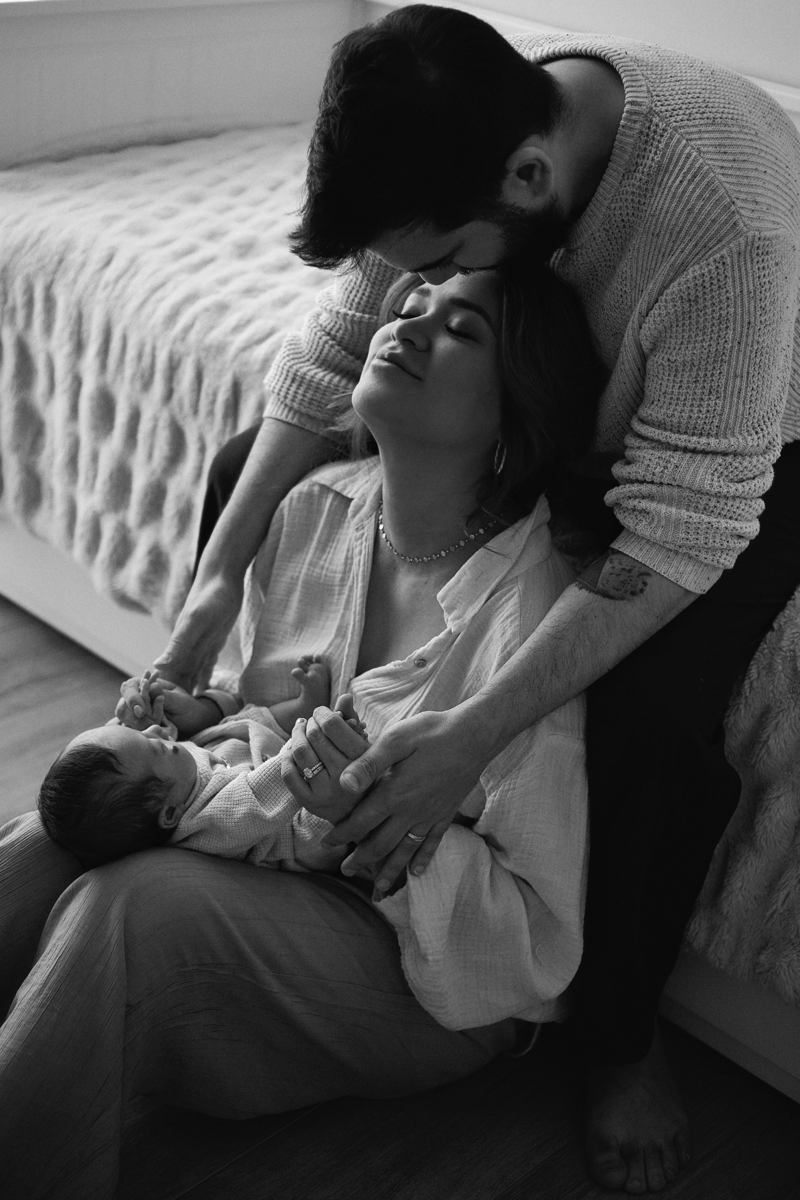 B&W: father kissing mother's head while she holds newborn on the floor