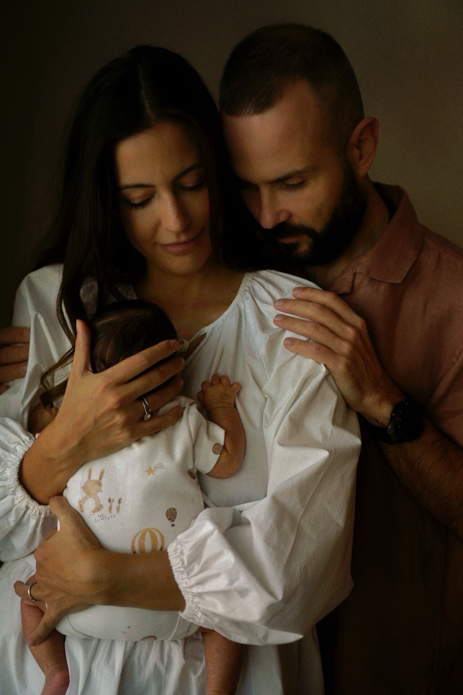 Parents holding newborn close in a dark intimate embrace