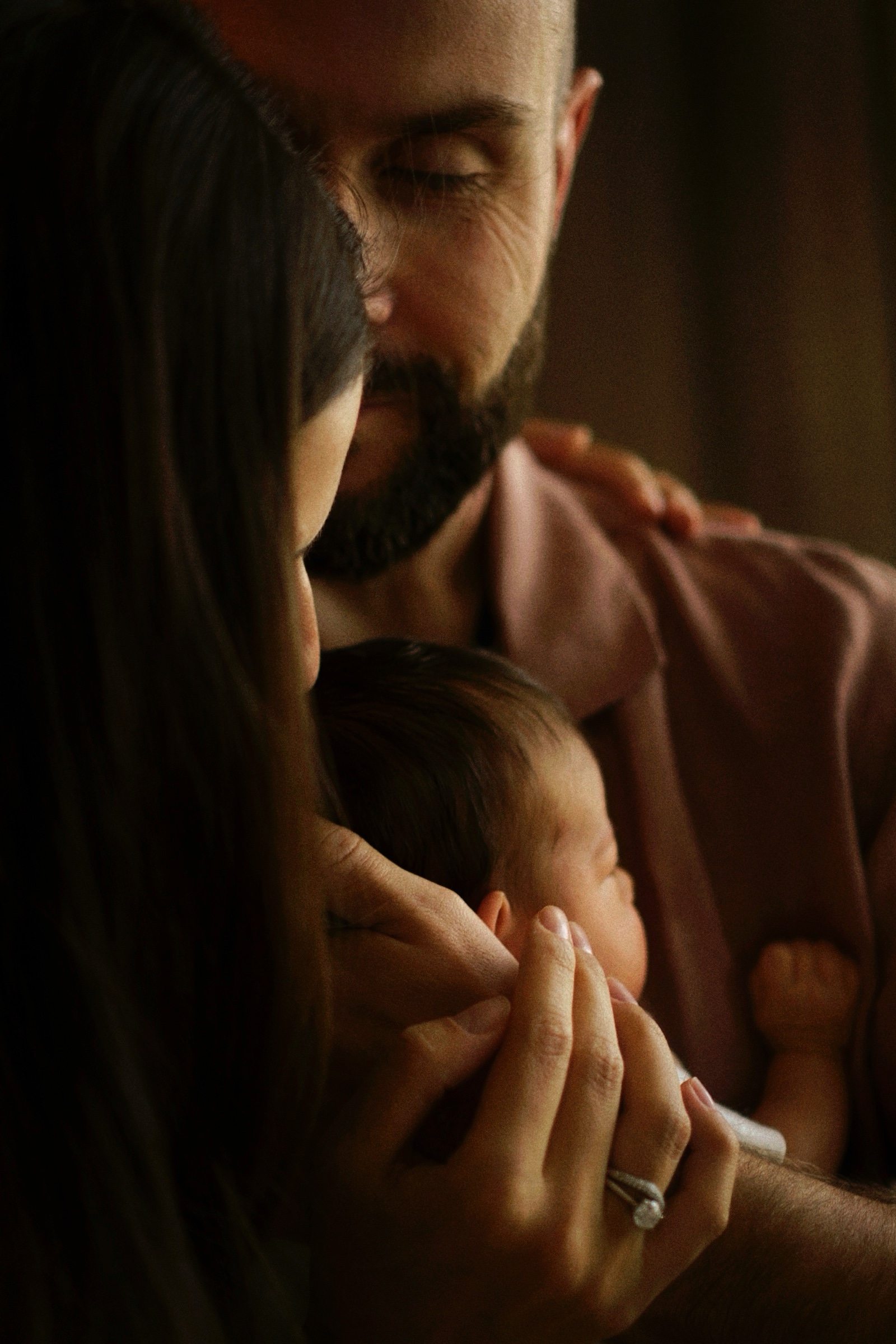 Dark moody close-up of father kissing mother as she cradles newborn