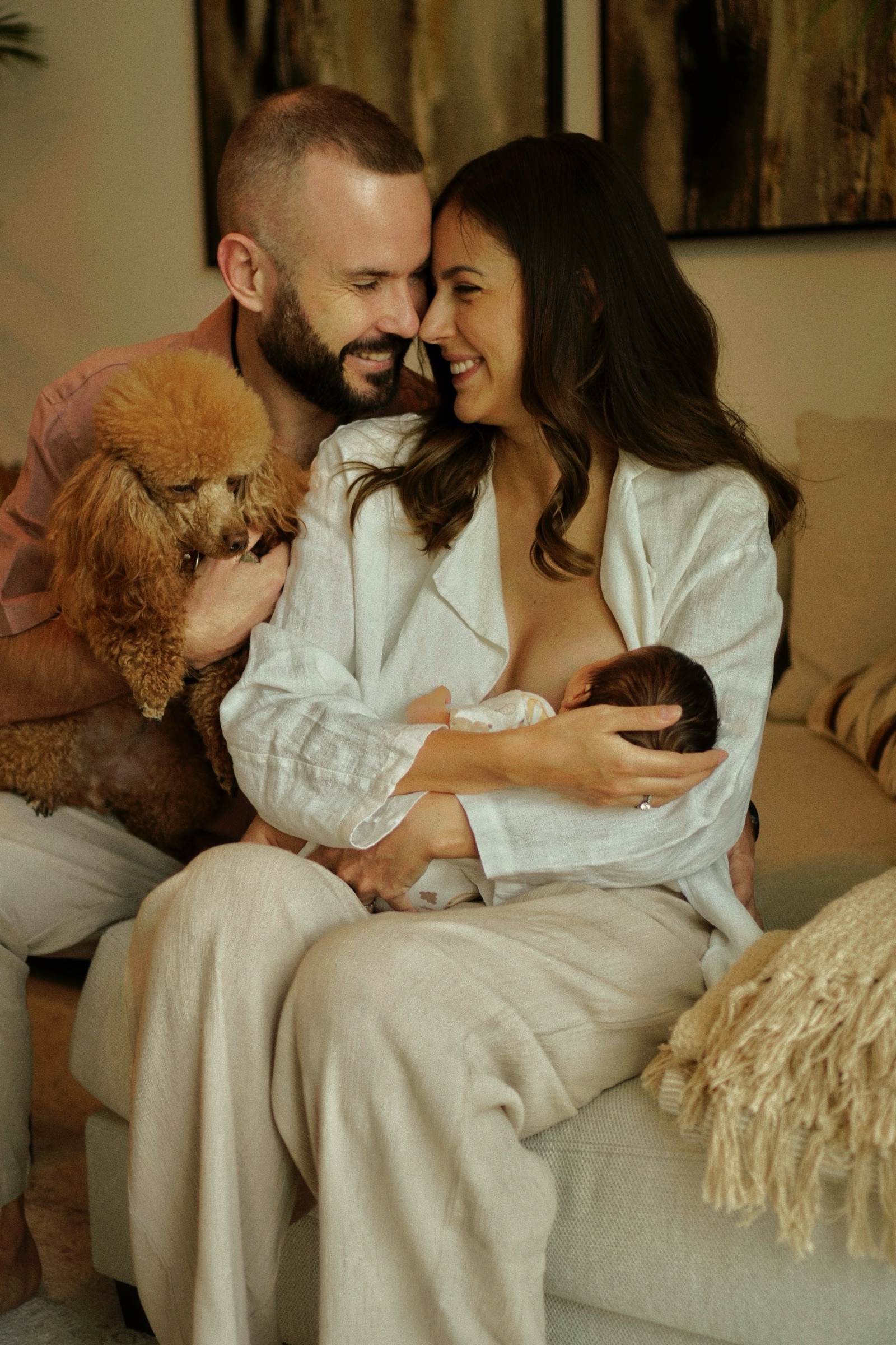Parents laughing with their golden poodle and nursing newborn on the sofa