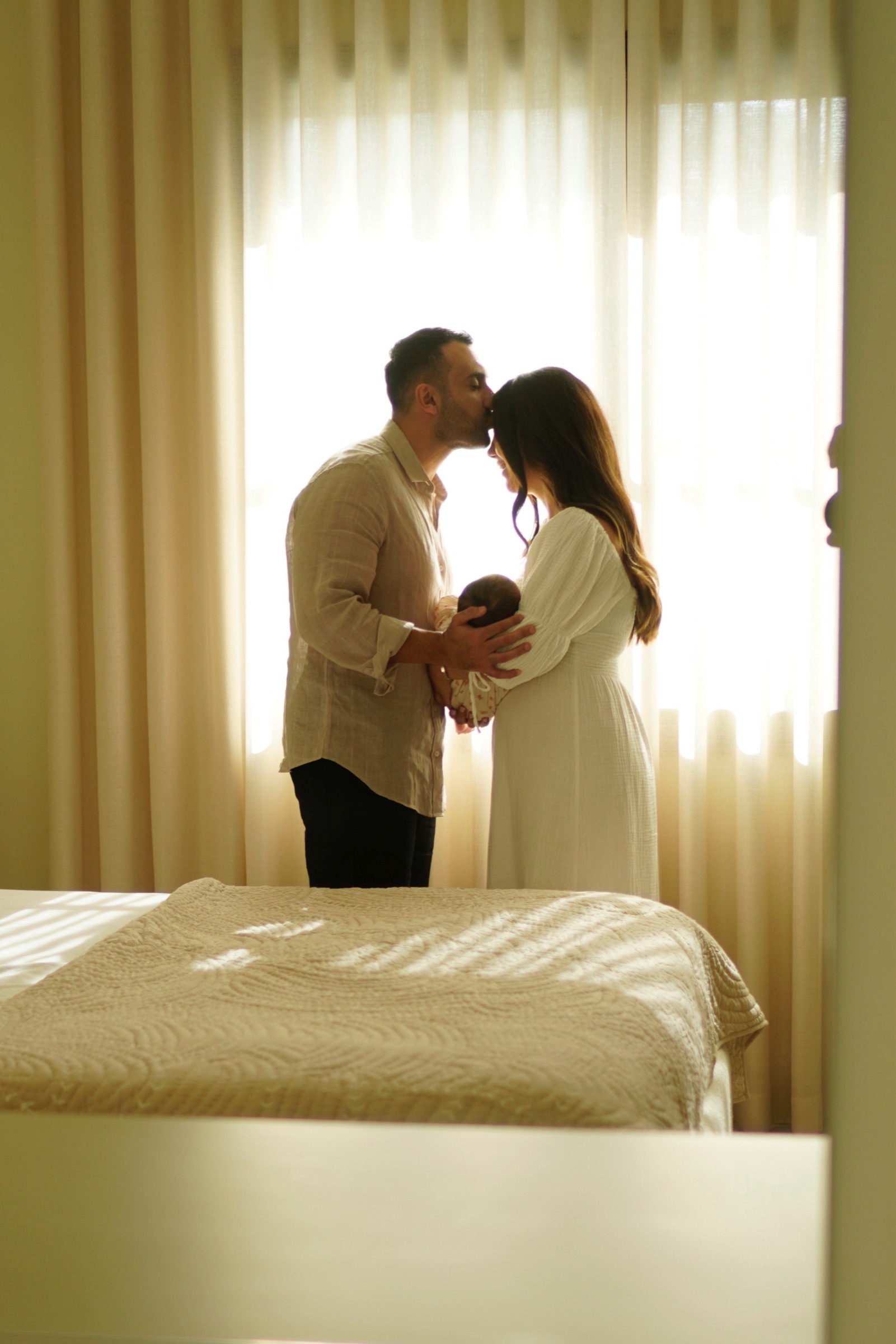 Parents kissing by the window holding their newborn in backlit silhouette