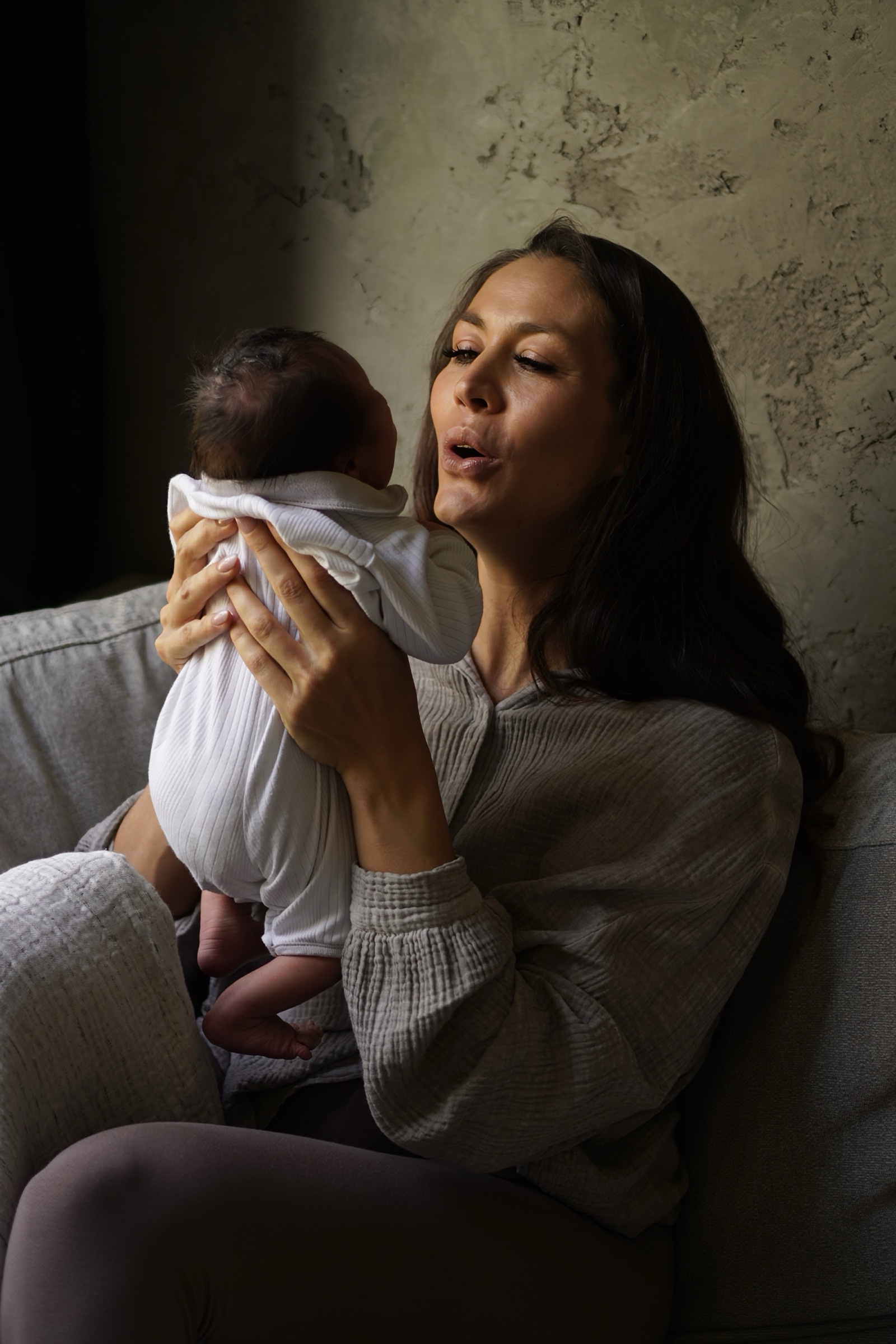 Mother in grey holding newborn up close, dramatic moody light against textured wall